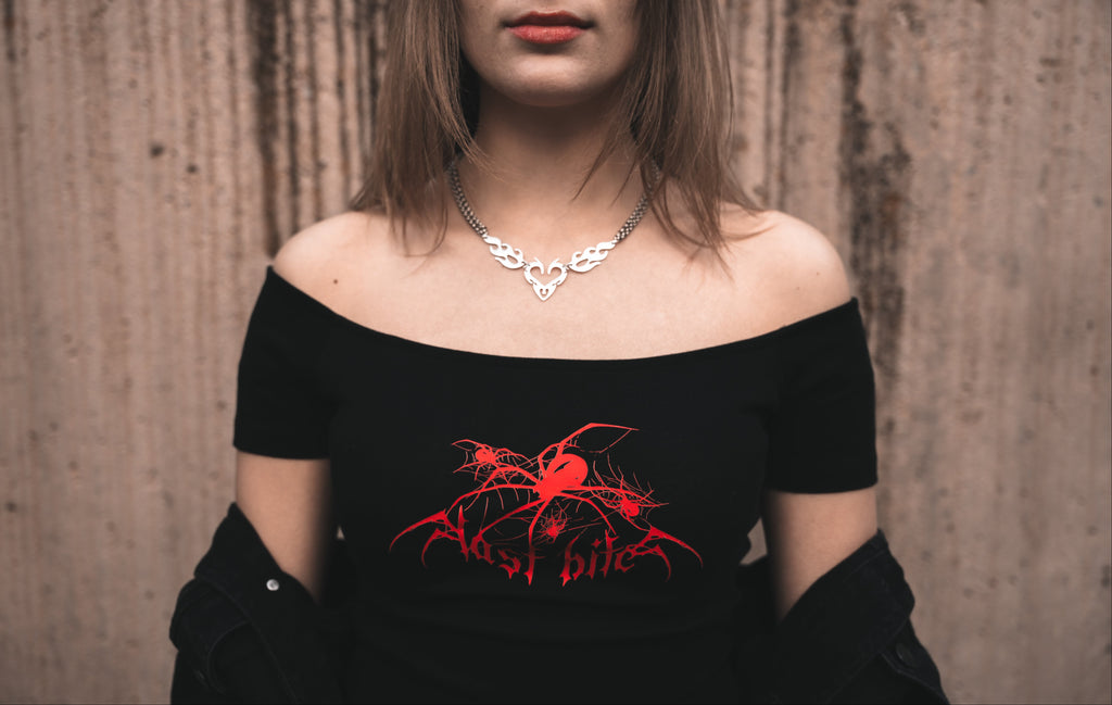 Model wearing a white off-the-shoulder black cotton red Last Bite crop top with a striking red abstract spider motif. Styled with a draped black jacket and silver tribal-style necklace.