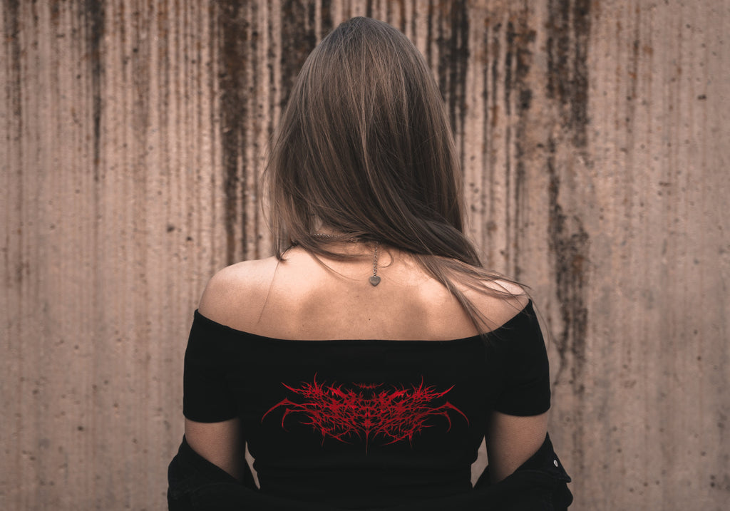 Model wearing a white off-the-shoulder cotton Last Bite crop top with a striking red abstract spider motif. Styled with a draped black jacket and silver tribal-style necklace.