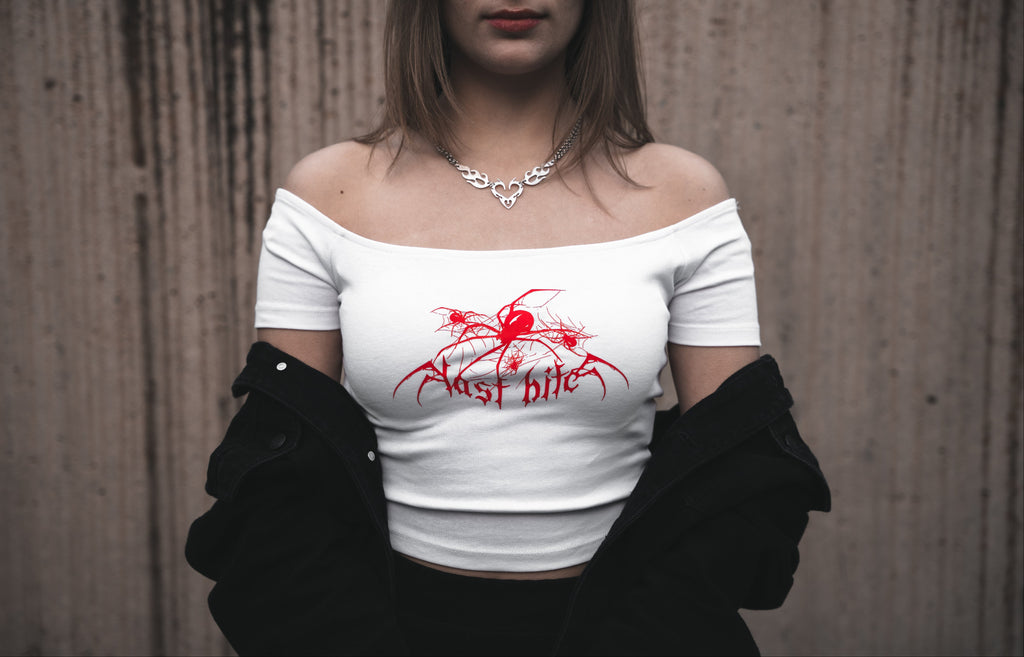 Model wearing a white off-the-shoulder cotton Last Bite crop top with a striking red abstract spider motif. Styled with a draped black jacket and silver tribal-style necklace.