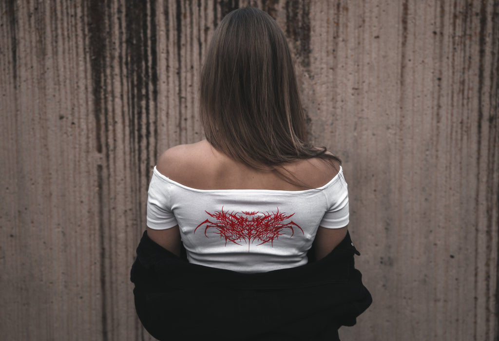 Model wearing a white off-the-shoulder cotton Last Bite crop top with a striking red abstract spider motif. Styled with a draped black jacket and silver tribal-style necklace.