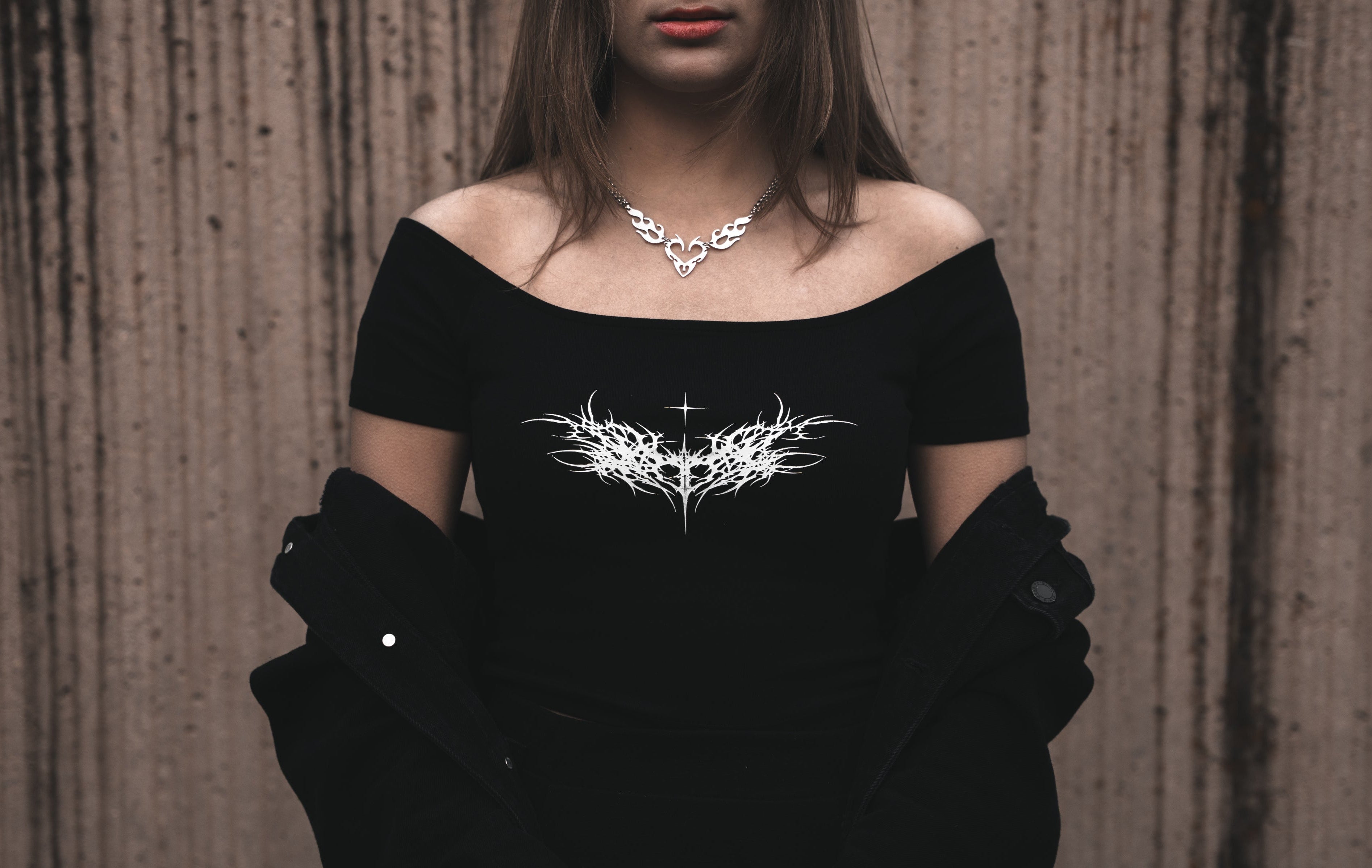 Model wearing a black off-the-shoulder cotton dissolved love crop top featuring a white neo-tribal heart and thorn graphic. Styled with a black jacket and a silver heart-shaped tribal necklace.