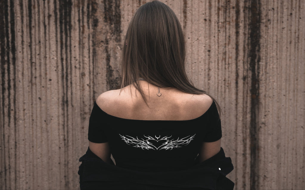 Model wearing a black off-the-shoulder cotton dissolved love crop top featuring a white neo-tribal heart and thorn graphic. Styled with a black jacket and a silver heart-shaped tribal necklace.

