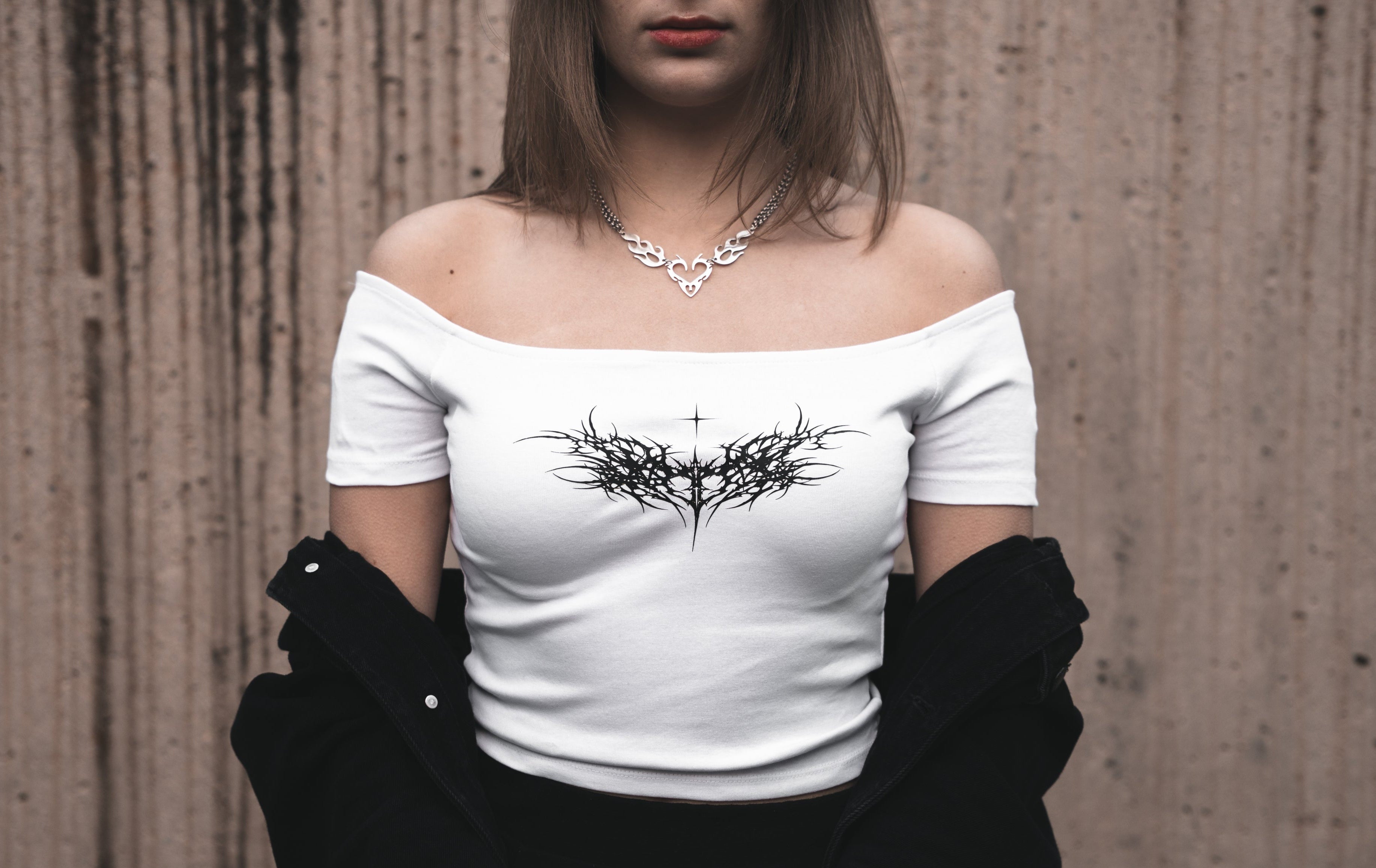 Model wearing a white off-the-shoulder cotton dissolved love crop top featuring a white neo-tribal heart and thorn graphic. Styled with a black jacket and a silver heart-shaped tribal necklace.