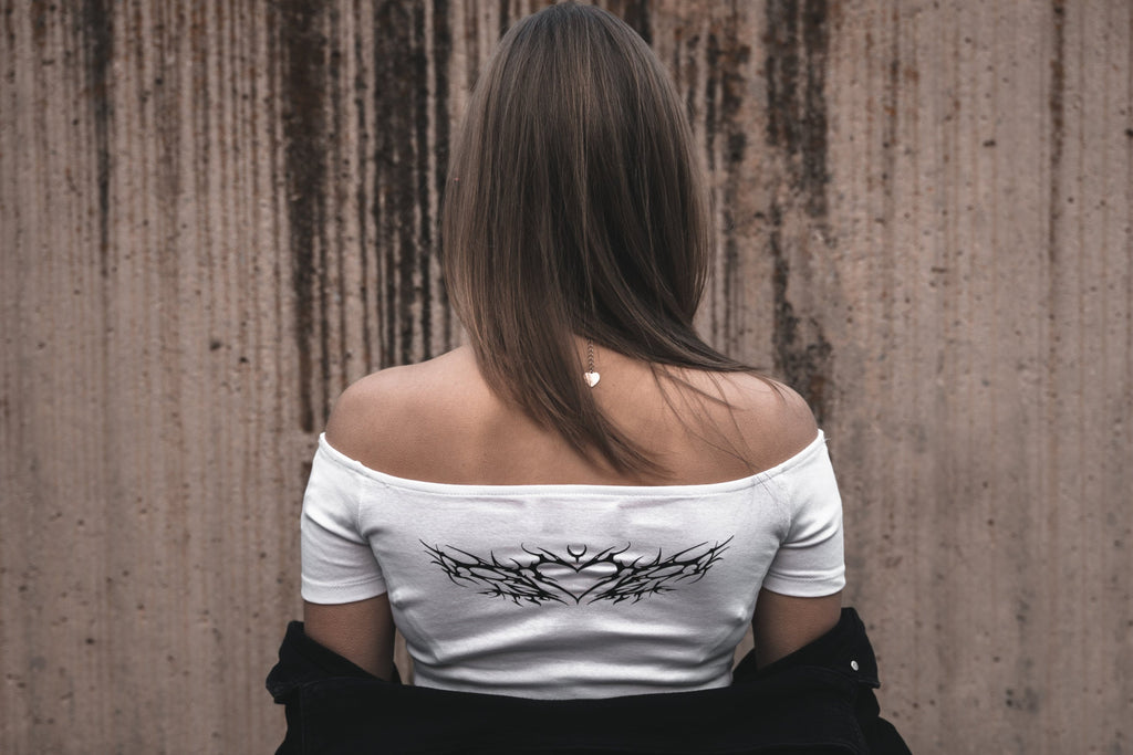 Model wearing a white dissolved love off-the-shoulder cotton dissolved love crop top featuring a white neo-tribal heart and thorn graphic. Styled with a black jacket and a silver heart-shaped tribal necklace.

